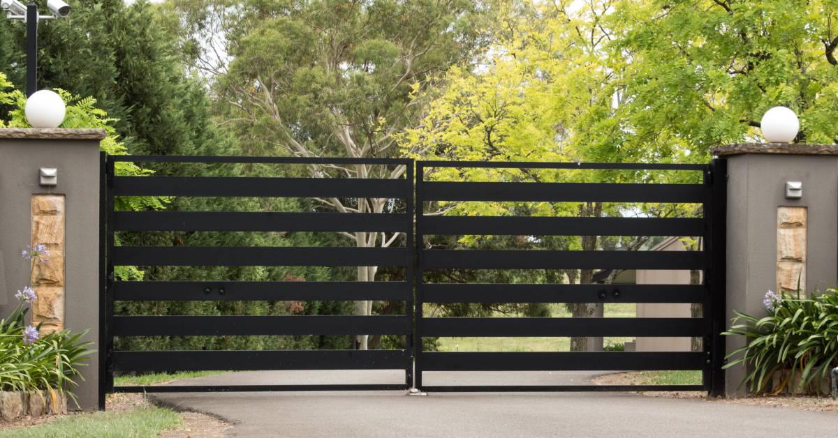 A black metal gate with two decorative concrete pillars securing a driveway. Past it, the drive continues through some trees.